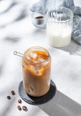 Ice latte coffee in a glass with milk on a light background with coffee beans and morning shadows.
