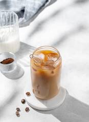 Ice latte coffee in a glass with milk on a light background with coffee beans and morning shadows.