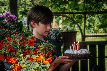 A boy holds a decorated strawberry cheesecake cake with chocolate on the edge. The 11th candle is lit on top of the cake, celebrating his 11th birthday. The background is softly blurred, highlighting 