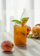 Ice tea with peach, mint in a glass on a white table near window with fresh fruits and shadow.