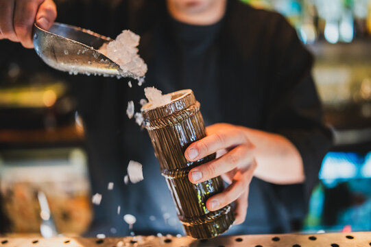 Close-up of a bartender scooping ice into a brown textured cocktail glass on a bar counter, capturing the drink preparation process in a nightlife setting.