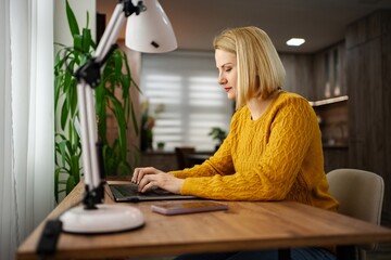 Focused woman in yellow sweater working on laptop at home desk with bright lamp and plants in a...