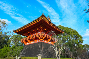 The brightly painted orange and white Kyozo, or sutra repository, at Ninna-ji Temple in Kyoto stands out with its unique architectural style against a backdrop of green trees and a clear blue sky.