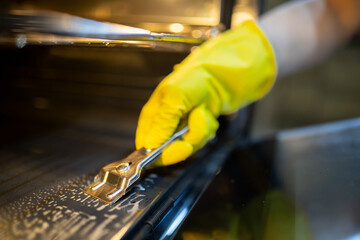 Cleaning the oven with care using a scraper tool and wearing rubber gloves in a well-lit kitchen