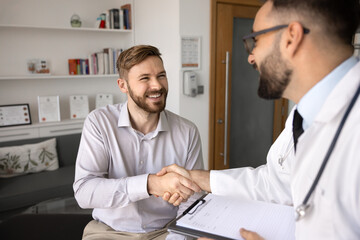 Cheerful young patient man giving handshake to Latin medical practitioner after examination in...