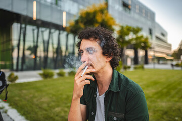 Man with curly hair sits on a park , smokes a cigar and rests