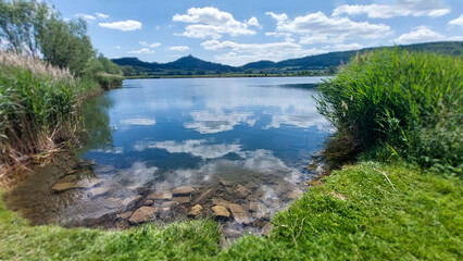 Calm lake reflecting clouds and mountains under a bright blue sky near a lush green shore