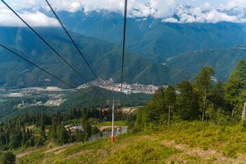 The red cab on the cable car stands against the backdrop of a summer forest and mountain peaks.