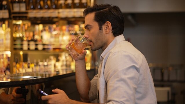 Smart man drinking cocktail in Old fashioned waiting friends or couple while chatting on smartphone to wait at counter bar with bartender preparing drinks at nightclub Friday meeting night. Vinosity. - Powered by Adobe
