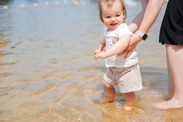 a joyful baby stands with supportive adult by the water's edge, experiencing the warmth of the sun...