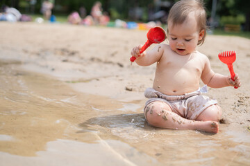 adorable child enthusiastically playing with red shovel and rake on sandy beachside with water...