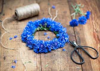 Handmade summer flower wreath with fresh deep blue cornflowers (Centaurea cyanus), styled on a rustic wooden backdrop. Decorative summer floral art handmade concept. Selective focus.