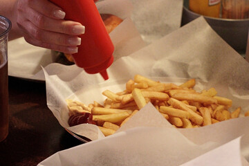 close up dinner scene showing hands pouring ketchup on french fries in a paper tray. Tasty fast...