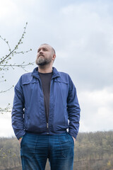 Man standing confidently outdoors in a blue jacket under a cloudy sky near a scenic landscape with rolling hills