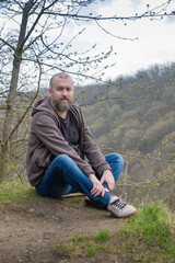 Man sitting on hillside in nature with trees and mountains in the background during cloudy day
