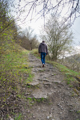 Hiker walks on rocky path through forest in early spring, surrounded by budding trees and cloudy sky