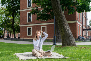 Exhausted sweaty woman drinking water from bottle while working on laptop sitting on blanket on...