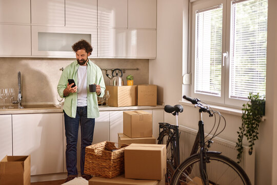 Man organizes his belongings in the kitchen of a new home while checking his phone for details about the move. Moving boxes and a bicycle are present in the space.