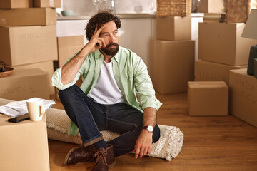 A man dressed casually sits on a rug surrounded by cardboard boxes, reflecting on his recent move....