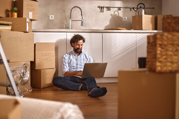 A man sits on the floor of a new kitchen surrounded by boxes, focused on his laptop as he organizes...