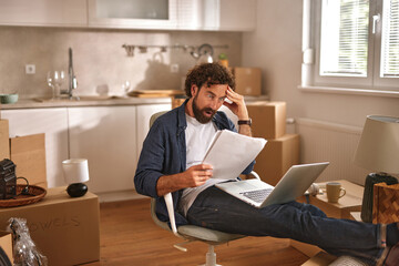 A person sits in a home office surrounded by moving boxes, looking stressed while reviewing...