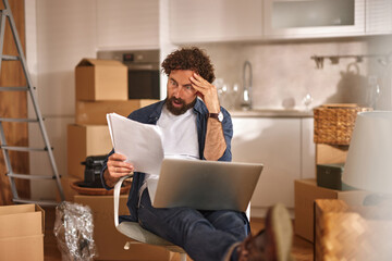 A man with curly hair is seated in a modern kitchen filled with unpacked boxes. He reviews important documents on his laptop, appearing deep in thought.