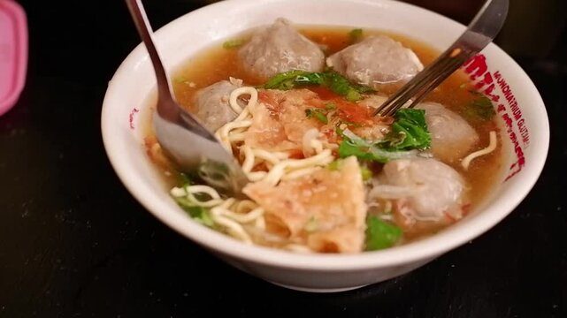 A hand stirs a bowl of Indonesian bakso meatball soup using a spoon and fork. The dish includes noodles, fried tofu, and meatballs served in hot broth on a wood-patterned table.
