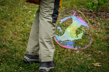 A child in a purple vest creates a large, colorful soap bubble using wands in a park setting,...