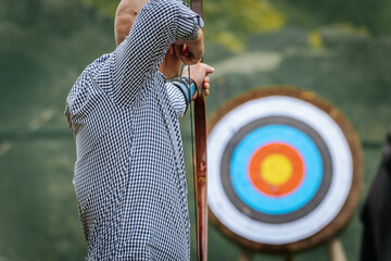 A bald man in a checkered shirt carefully aims a bow at a colorful archery target in an outdoor...