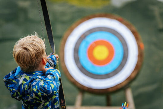 A young boy aims a bow at a colorful archery target, focusing intently while holding the string back, wearing a vibrant blue patterned jacket.