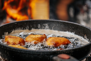 A black skillet with bubbling oil and golden pastries fries over hot coals, with a slotted spatula stirring the food in an outdoor rustic setting.