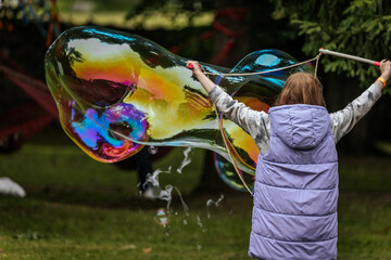 A child in a purple vest creates a large, colorful soap bubble using wands in a park setting,...