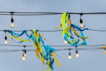 Blue, yellow, and turquoise ribbons hang alongside glowing string lights on wires, creating a...