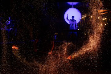 A fire performer creates a fiery display with sparks flying, while a drummer plays under a glowing...