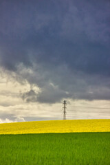 Minimalistic vertical landscape of yellow rapeseed fields blossoming under a storm and with an electric tower in the middle. Three colour stripes.