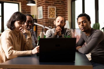Focused diverse group watching crime or investigative documentary on laptop. Multiethnic friends seated around table, visibly intrigued by movie on digital device, indoor hangout.