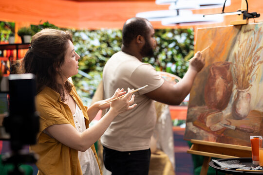 Woman painter filming her colleague during his creative process and explaining the painting technique for online tutorial. Diverse artists creating virtual classroom to teach art. Digital education.