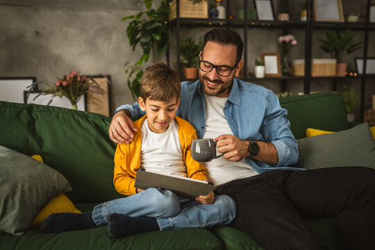 Father hold cup of coffee and look son while he use tablet for fun