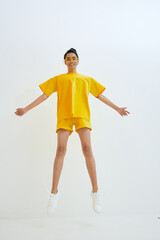 Woman jumping in yellow casual outfit against white background, wearing sunglasses and sneakers,...