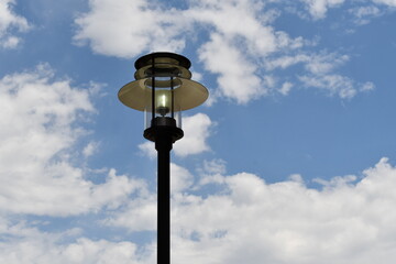 Lamp post turned on during the daytime. Beautiful fluffy clouds in the background.