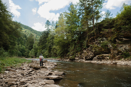 Fisherman walking on rocks in river surrounded by lush green forest - Powered by Adobe