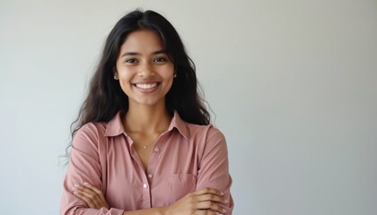 Young Indian woman with crossed arms, smiling brightly at camera. Long dark hair, wears pink button-up shirt. Portrait shot in studio with clean white backdrop, conveying confidence, positivity.