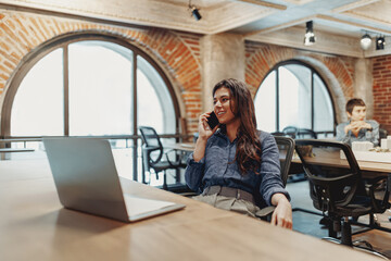 A young woman is making an important phone call while also working on her laptop in a contemporary office environment