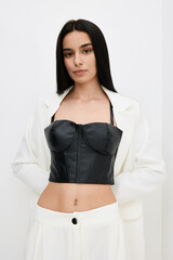 young woman wearing a black leather corset and white blazer standing against a plain white...