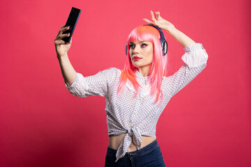Caucasian woman wearing wireless headphones and taking playful selfies against vibrant pink...