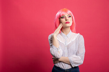 Young female individual with hand on cheek, looking upward in thoughtful contemplation. Pensive woman with pink hair stands against isolated background, appearing focused and reflective.