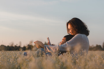 Happy mother hugging her son while sitting together in flower field at golden hour sunset. Enjoy relax family moment outdoor using smartphone. Love, motherhood, lifestyle, child, summer, Mother’s Day.