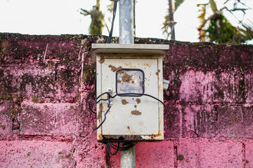A weathered electrical box stands against an old pink wall, showing signs of aging and exposure....