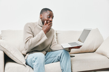 Happy young Black man with surprised expression, interacting with laptop on cozy couch, modern...