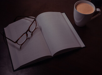 Tortoise-shell glasses, coffee cup, and open book arranged on a wooden desk